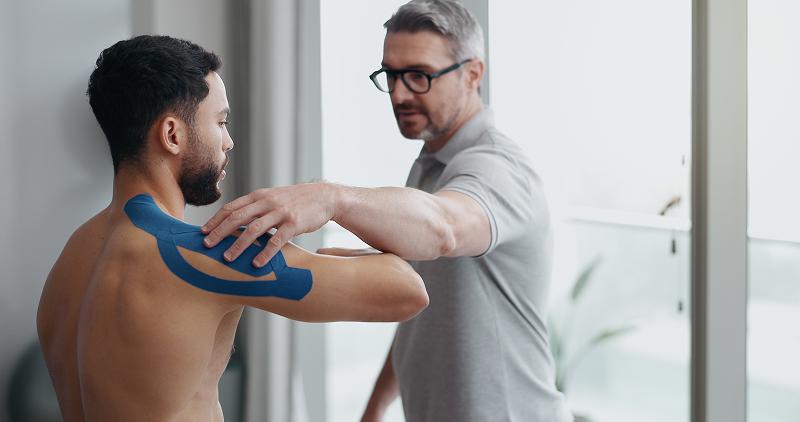 A healthcare professional applies blue therapeutic tape to a shirtless patient's shoulder during a medical examination.